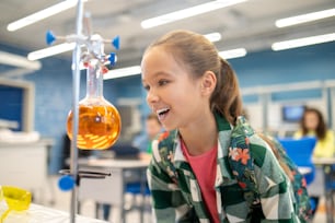 Delight. Smiling girl standing sideways to camera with delight looking at flask with colored liquid in chemistry class