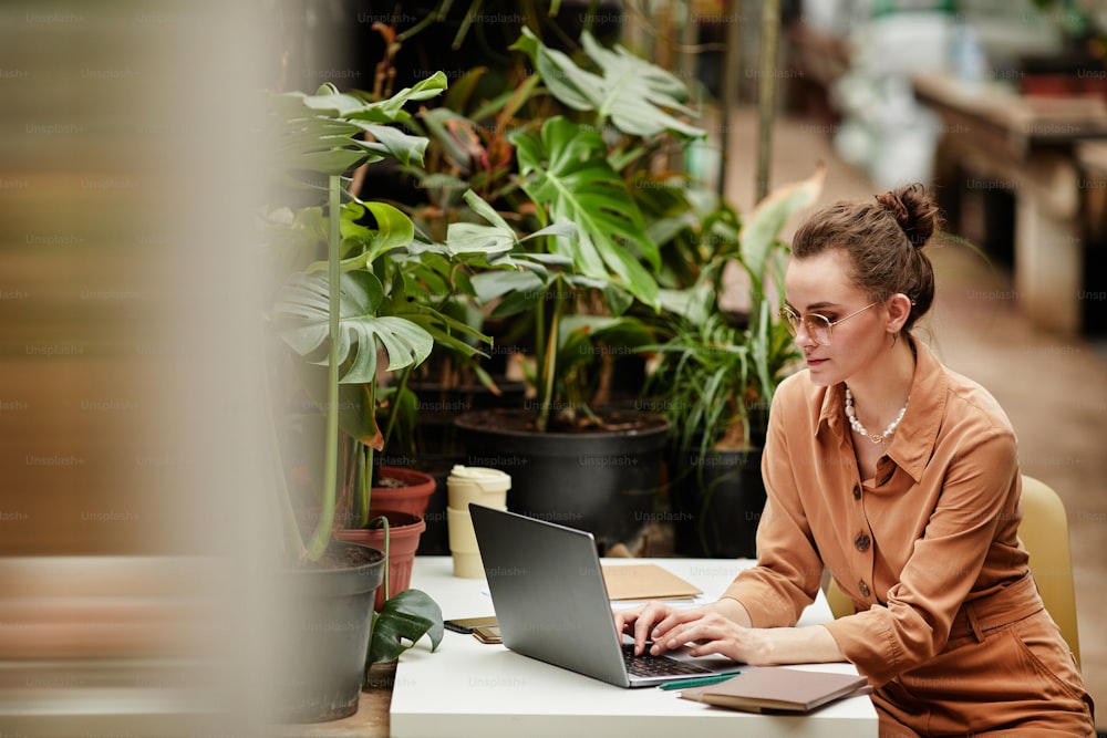 Jeune designer contemporaine ou propriétaire d’une petite entreprise travaillant sur Internet tout en étant assise près d’un bureau devant un ordinateur portable dans un magasin de fleuriste