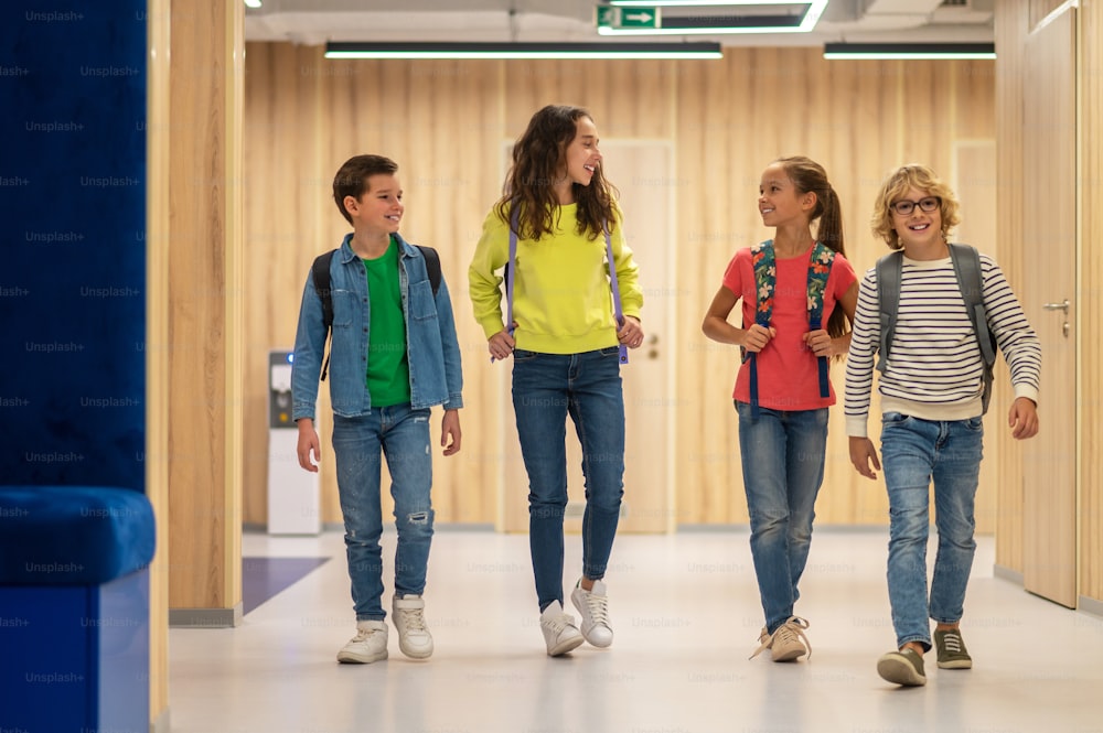 Horario escolar. Niñas y niños con mochilas en ropa casual charlando alegremente caminando por el pasillo iluminado de la escuela