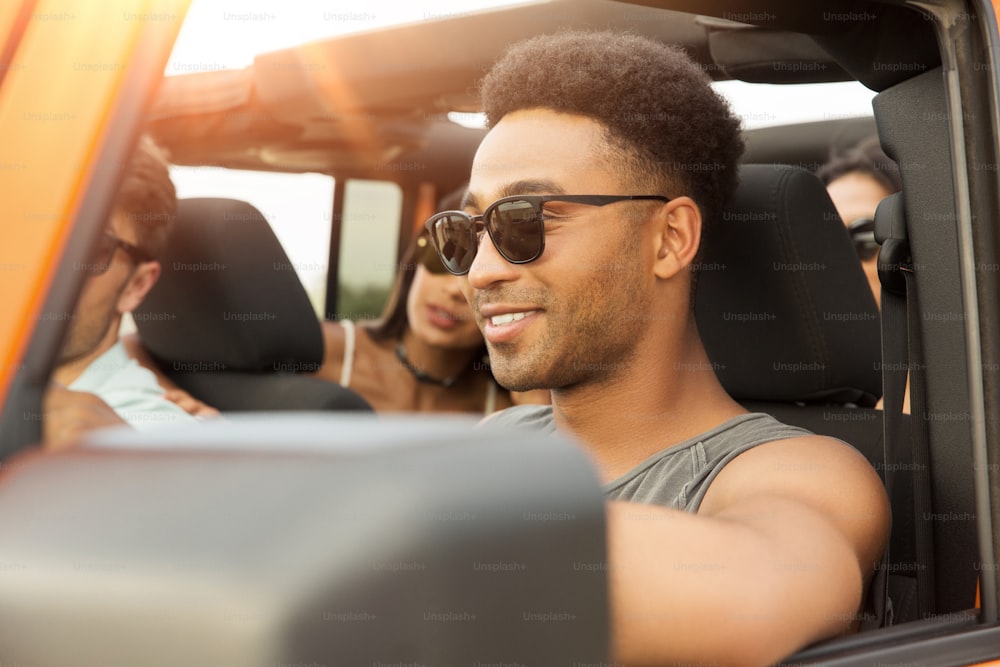 Joven africano sonriente conduciendo un coche y divirtiéndose con sus amigos