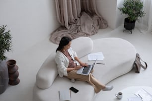 Young Asian businesswoman with lapto pon her knees concentrating on network while sitting on comfortable white sofa at home