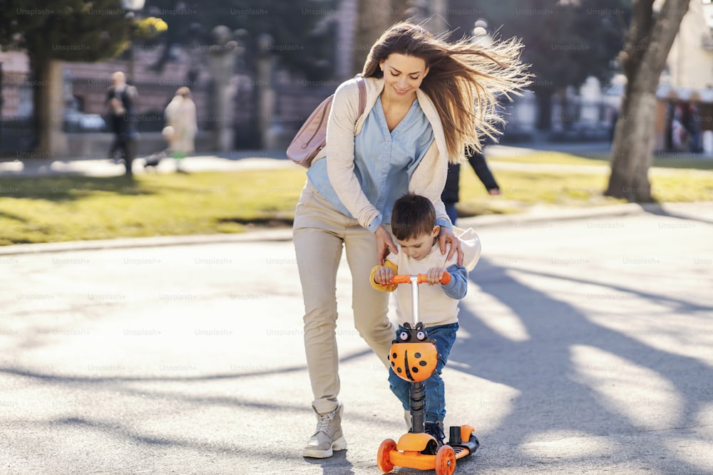 A mother pushing her son on a scooter in a park to go faster.