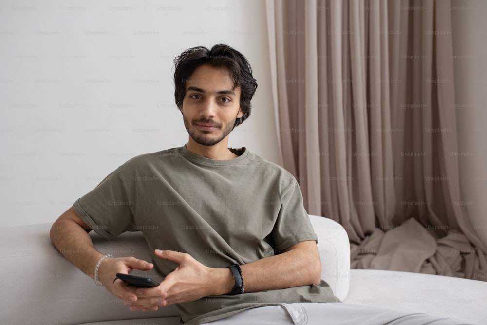 Young modern male manager with mobile phone sitting on white sofa in living room and communicating in online chat