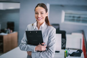 Porträt einer glücklichen Geschäftsfrau mit Klemmbrett in den Händen am Arbeitsplatz.