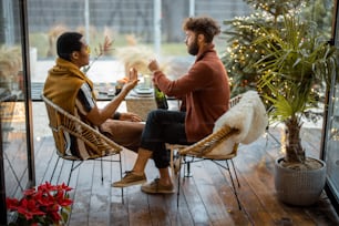 Two multiracial male friends drinking spakling wine, celebrating winter holidays, sitting by a dinner table at backyard with Christmass tree. Caucasian and hispanic man having a festive dinner