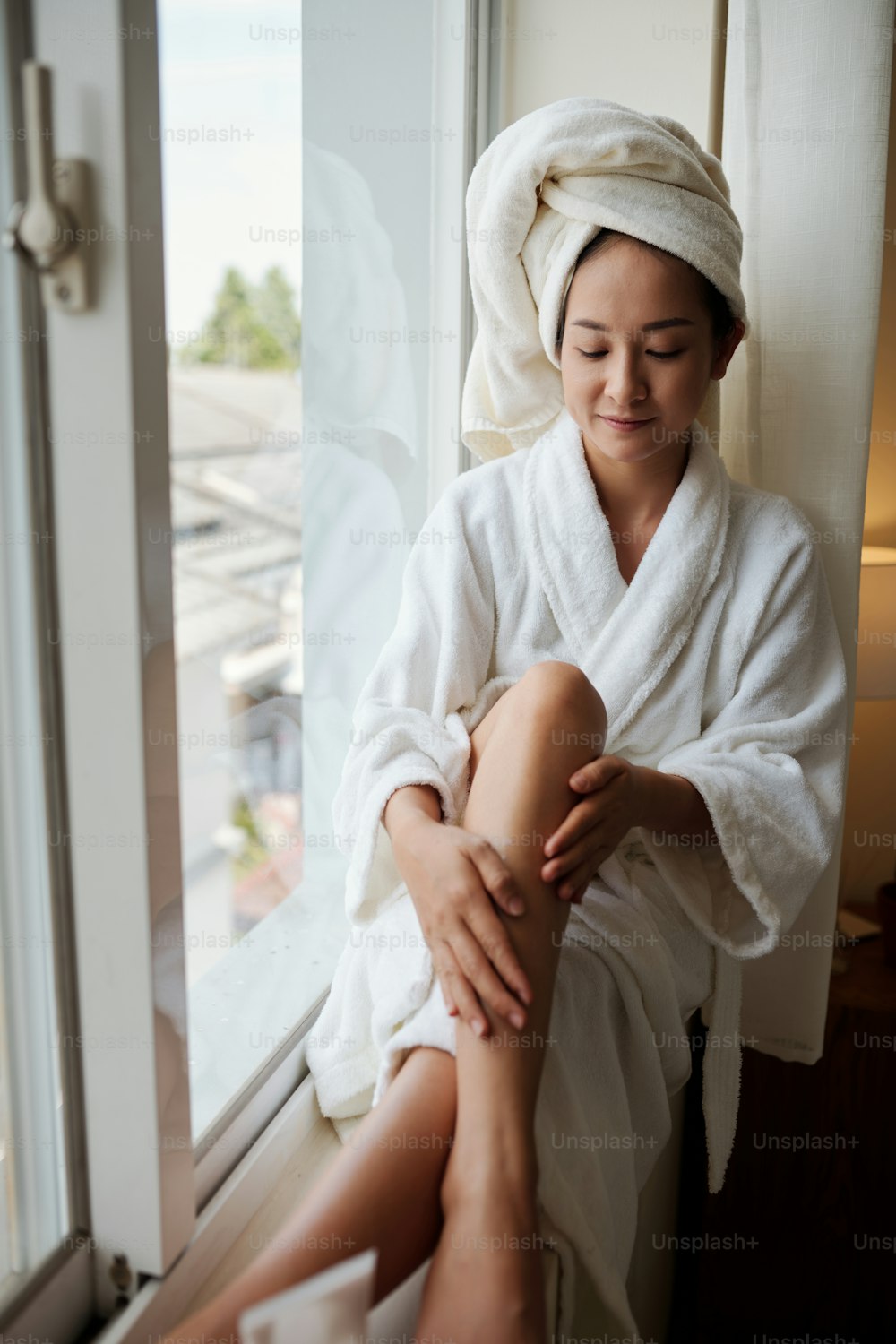 Mujer joven sonriente con bata de baño suave sentada en el alféizar de la ventana y aplicando loción corporal reafirmante