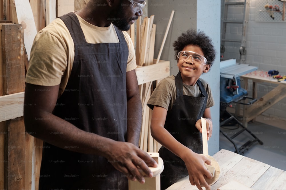 Retrato do menino afro-americano sorridente olhando para o pai enquanto desfrutam do tempo juntos na oficina de carpintaria