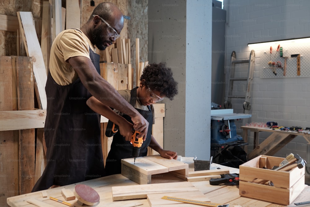 Ritratto di padre e figlio afro-americano che si legano in laboratorio mentre costruiscono mobili in legno insieme