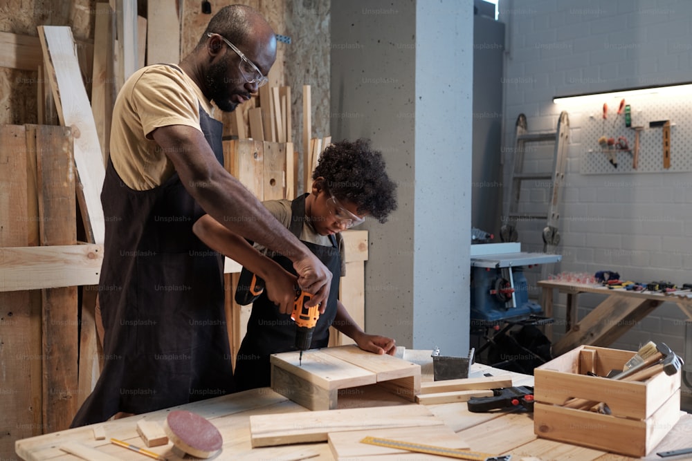 Porträt eines afroamerikanischen Vaters und Sohnes, die sich in der Werkstatt verbinden, während sie gemeinsam Holzmöbel bauen