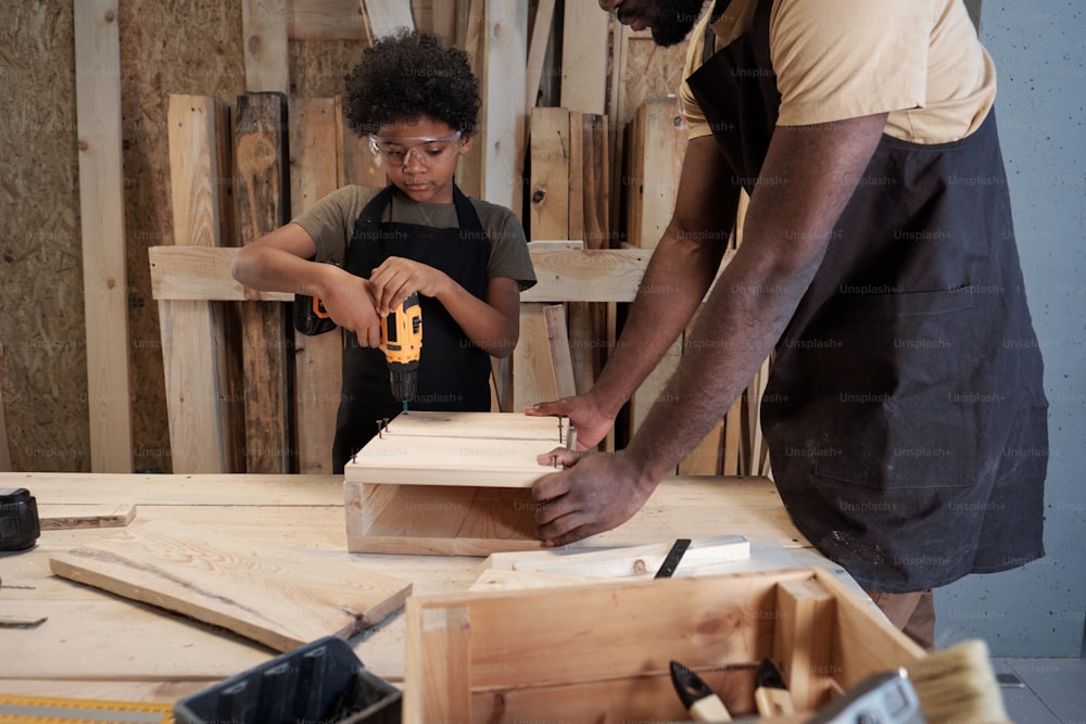 Retrato do menino afro-americano bonito construindo casa de pássaro de madeira enquanto se liga com o pai na oficina
