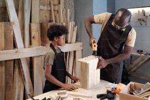 Ritratto in vita di padre e figlio afro-americani che lavorano insieme nel laboratorio di falegnameria e costruiscono mobili in legno