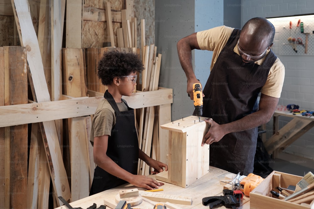 Retrato de cintura para arriba de padre e hijo afroamericanos trabajando juntos en un taller de carpintería y construyendo muebles de madera