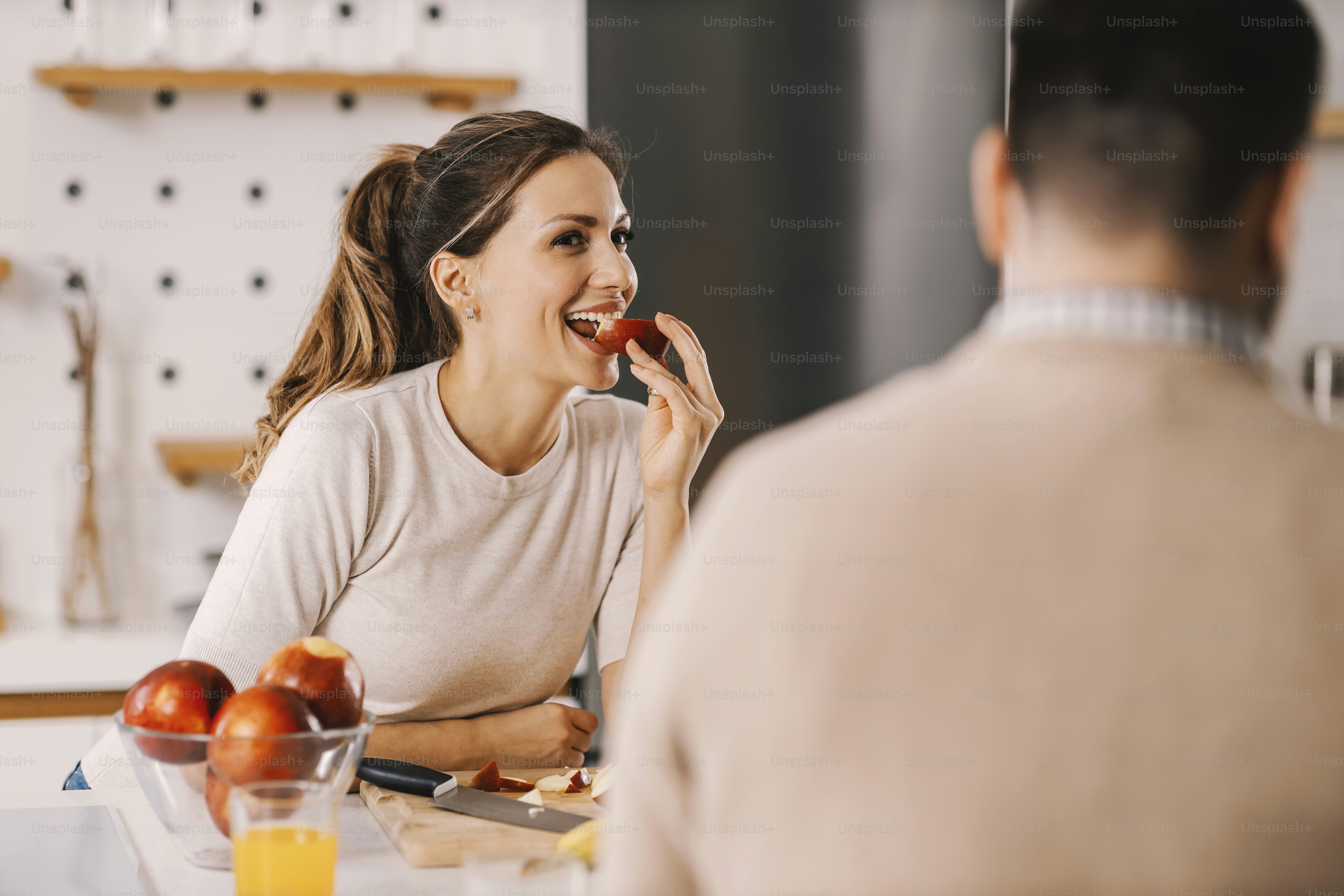 A wife eating slice of apple in kitchen and looking at husband.
