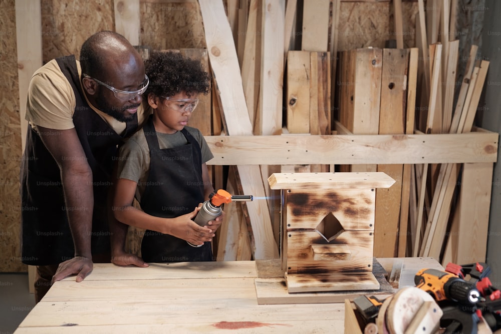 Porträt eines afroamerikanischen Vaters und Sohnes, die gemeinsam in einer Werkstatt ein hölzernes Vogelhaus bauen