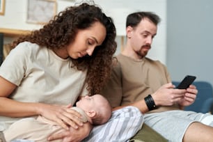 Joven mujer bonita mirando al bebé dormido en sus manos mientras está sentada en la cama contra su esposo con un teléfono inteligente por la mañana