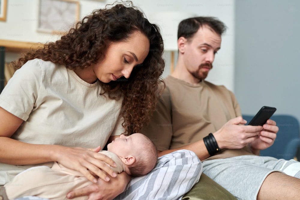 Young pretty female looking at sleeping baby on her hands while sitting on bed against her husband with smartphone in the morning