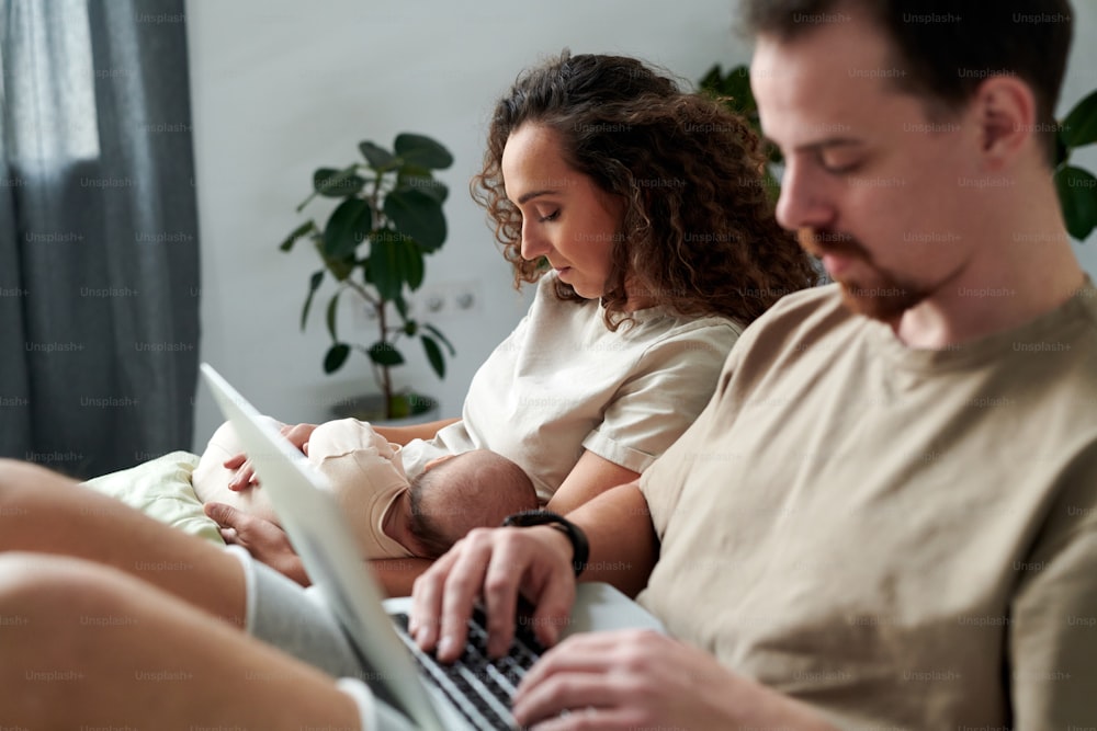 Young serene woman with newborn baby son on hands sitting on bed by her husband typing on laptop keyboard while networking