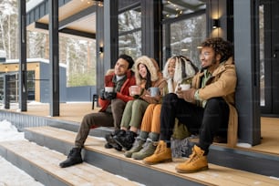 カントリーハウスの外の階段に座ってコーヒーを飲み、冬を楽しむ若い友人のグループ