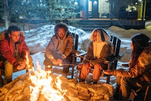 Grupo de amigos felices sentados en sillas y cocinando malvaviscos cerca del fuego durante sus vacaciones de invierno en la casa de campo