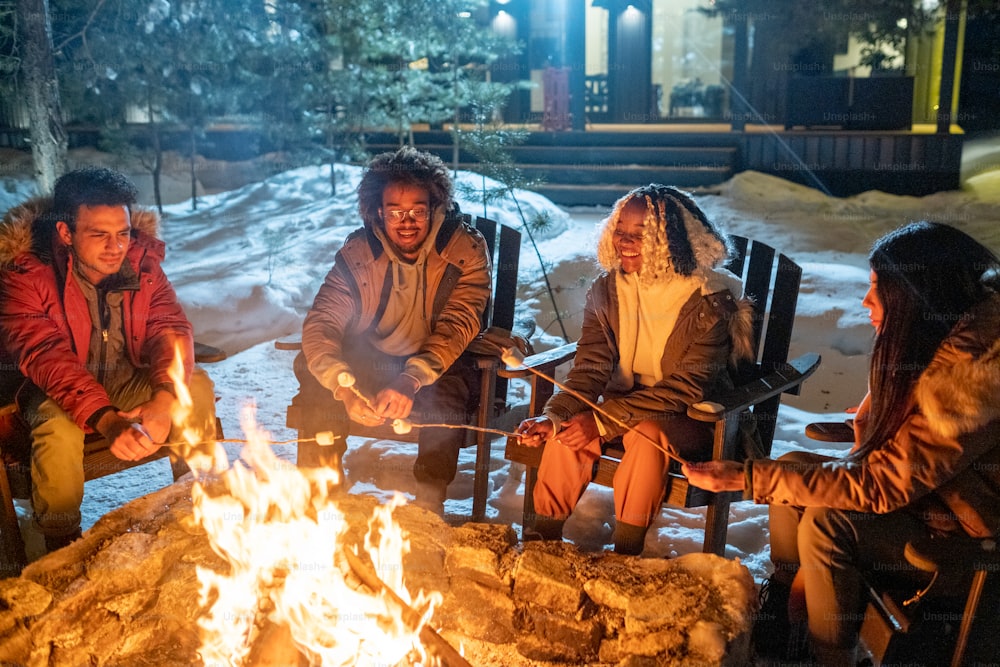 Gruppe glücklicher Freunde, die während ihres Winterurlaubs im Landhaus auf Stühlen sitzen und Marshmallows in der Nähe des Feuers kochen