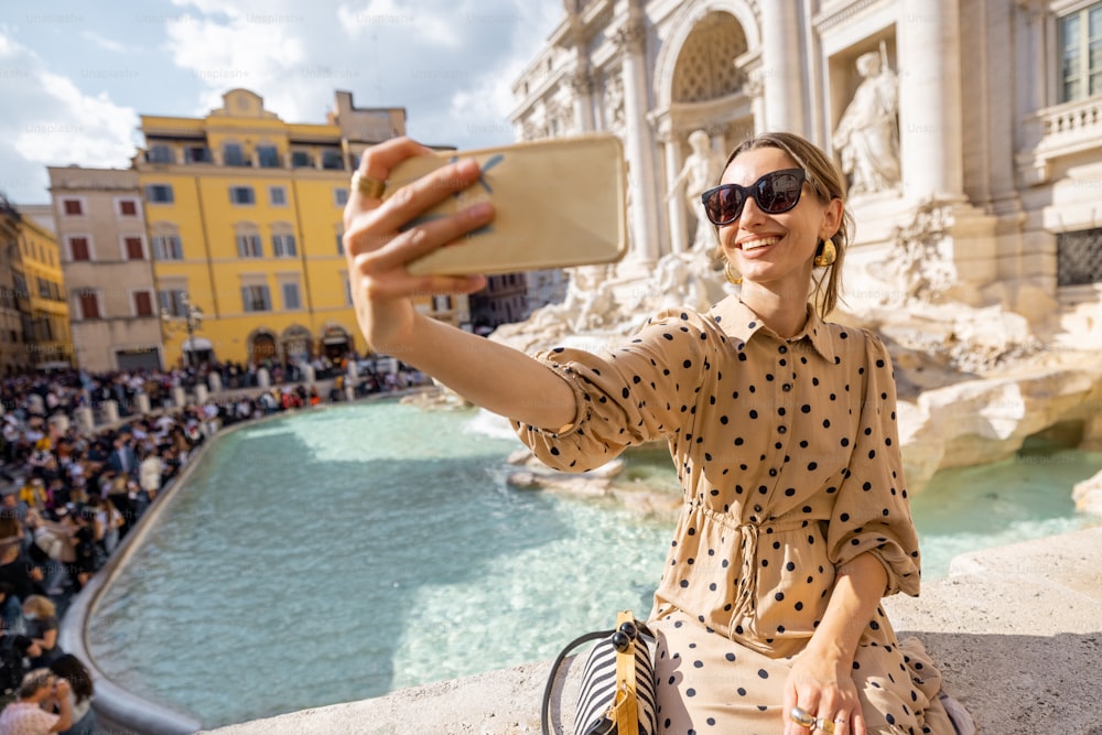 Woman taking selfie photo on background of famous di Trevi fountain in Rome. Traveling Italy on a summer day concept. Female traveler visiting italian landmarks