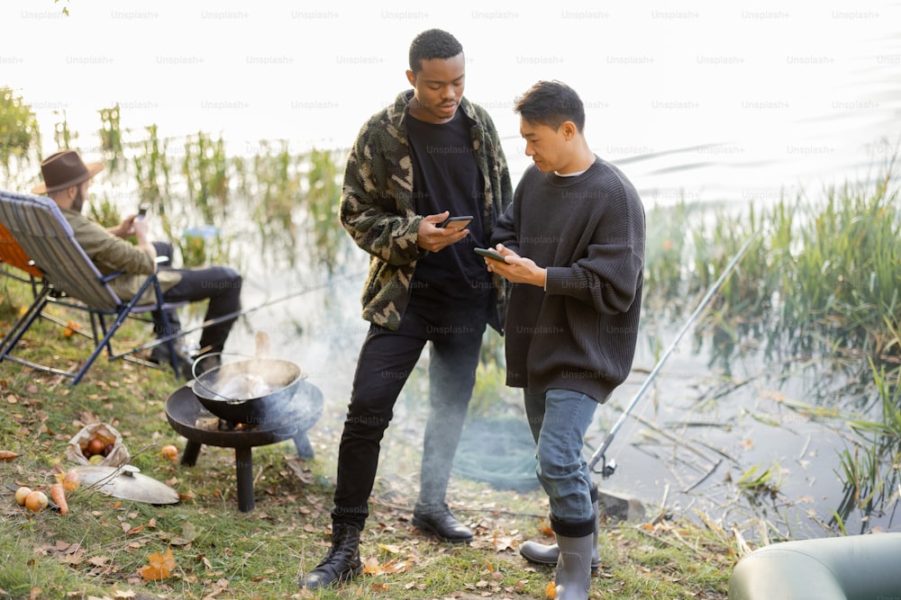 Asian and latin men using smartphones during resting on river or pond coast. Men fishing and cooking on river coast at autumn. Leisure and weekend in nature. Idea of friendship and spend time together