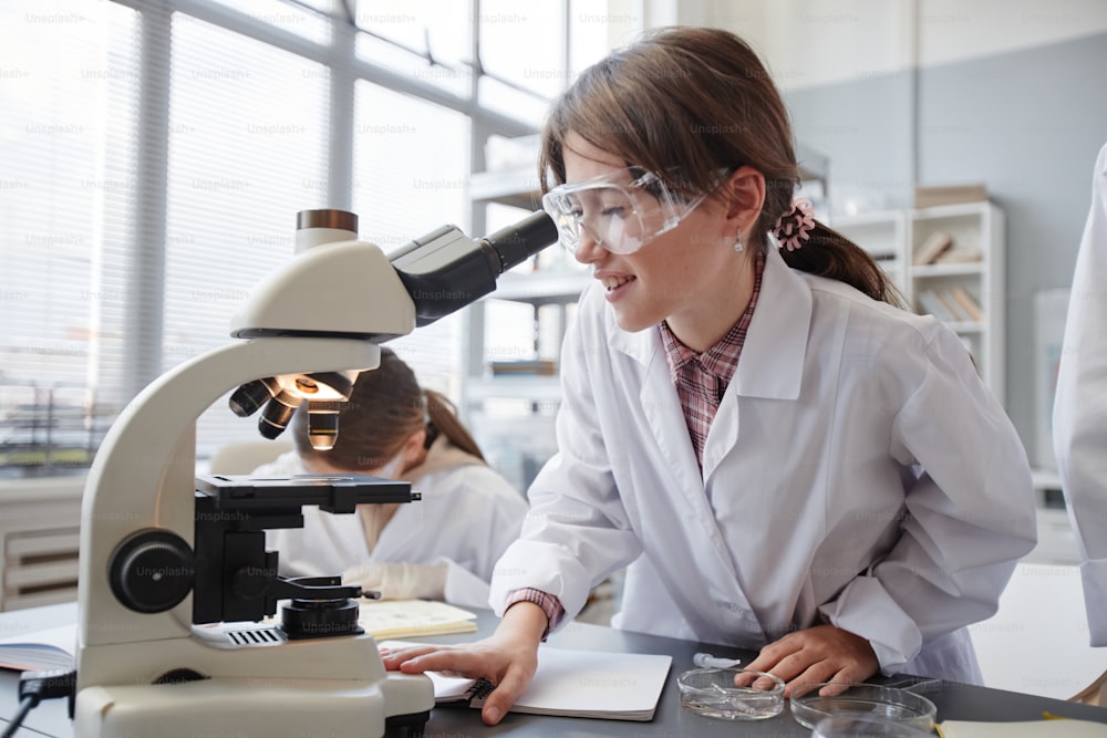 Retrato de vista lateral de una niña sonriente mirando al microscopio mientras disfruta de experimentos en el laboratorio de química de la escuela