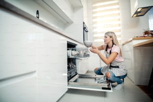 A woman in kitchen at home puts dishes into a dish washing machine.