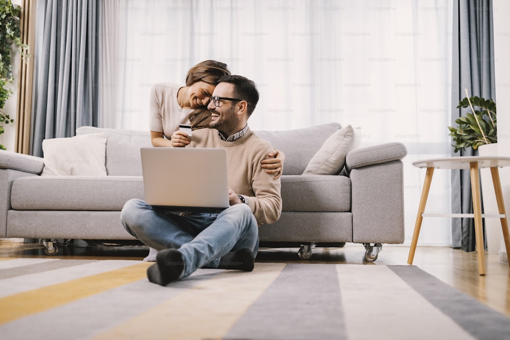 A couple in love embraces and uses a credit card for e-shopping at home.