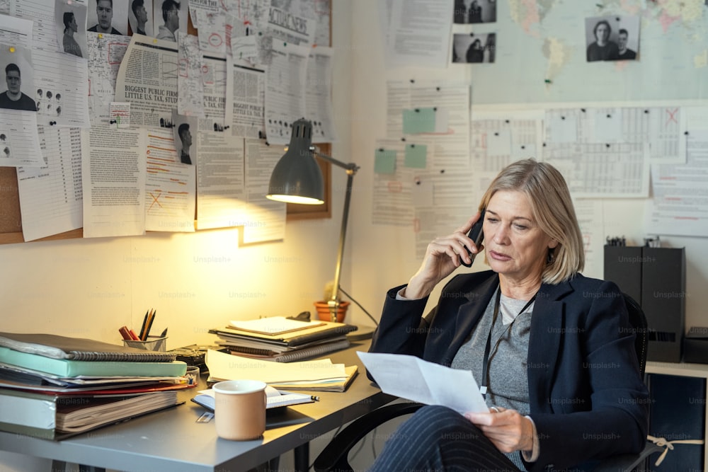 Mature female investigator questioning witness or victim on the phone while sitting by workplace and looking through case points