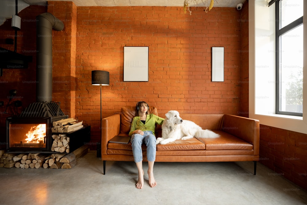 Junge Frau entspannt sich und hört die Musik, während sie mit ihrem süßen weißen Hund auf einer Couch zu Hause sitzt. Weitblick auf modernes Wohnzimmer im Loft-Stil mit Kamin