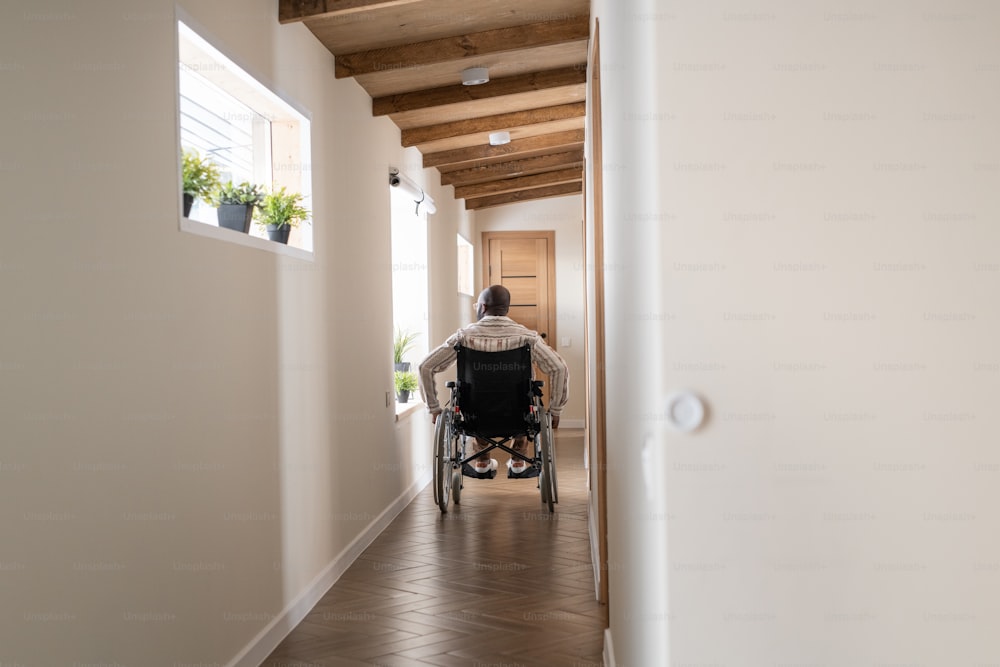 Vista traseira do homem com paralisia movendo-se em cadeira de rodas ao longo do corredor do grande apartamento cotemporário em direção à porta de entrada