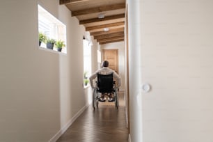 Back view of man with paralysis moving on wheelchair along corridor of large cotemporary apartment towards entrance door