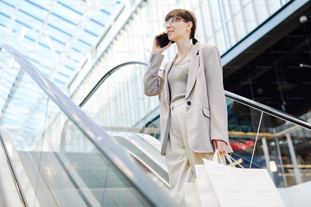 Retrato com vista lateral de jovem empresária falando no smartphone em shopping center enquanto está em pé na escada rolante e segurando sacolas