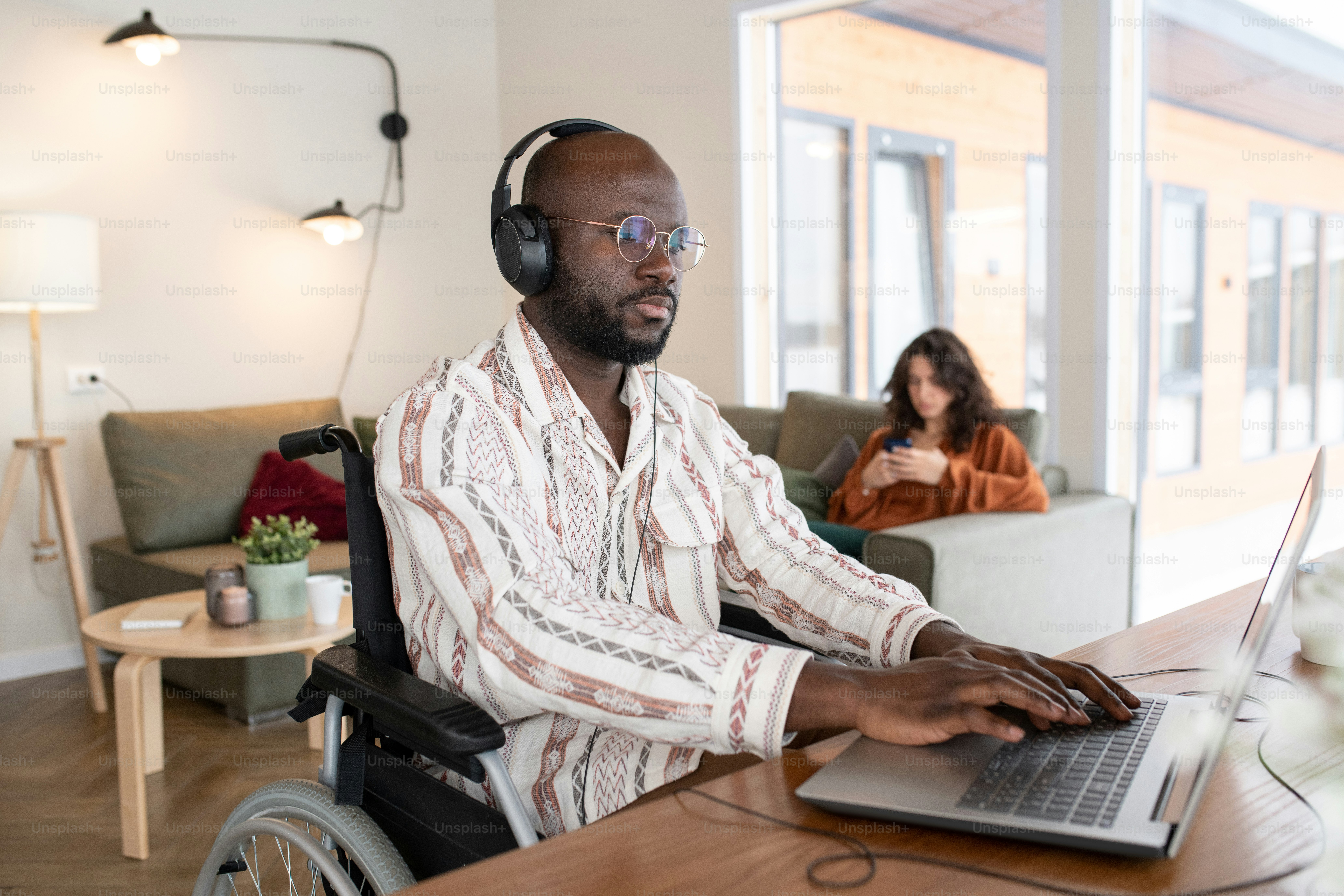 Hombre de negocios afroamericano serio con auriculares escribiendo junto a la mesa en la sala de estar mientras trabaja con datos en línea contra una mujer joven