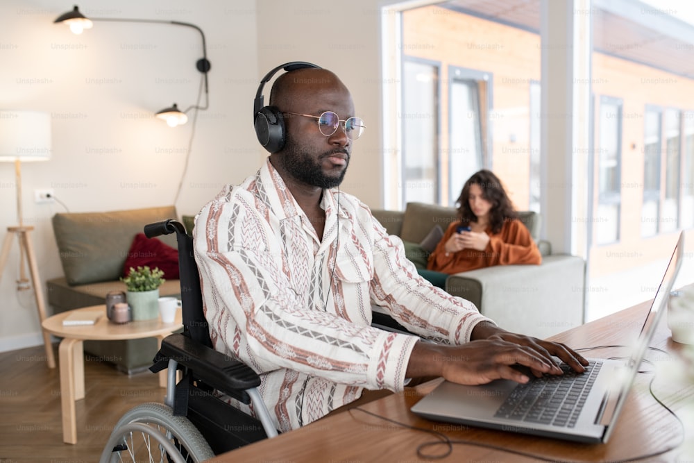 Ernsthafter afroamerikanischer Geschäftsmann in Kopfhörern, der am Tisch im Wohnzimmer tippt, während er mit Online-Daten gegen eine junge Frau arbeitet