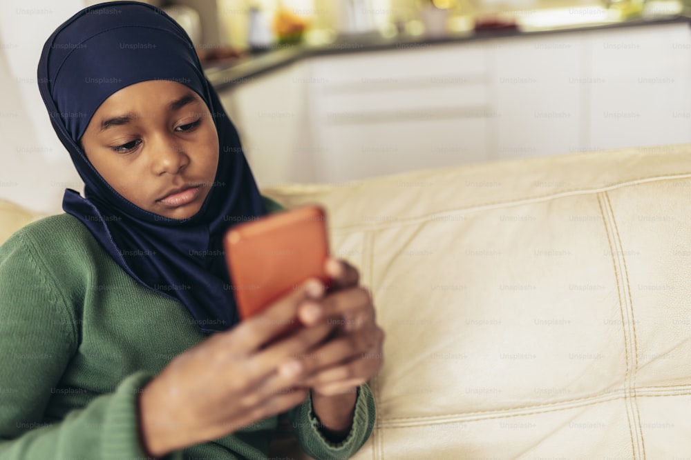 Muslim girl sitting on sofa at home using phone.