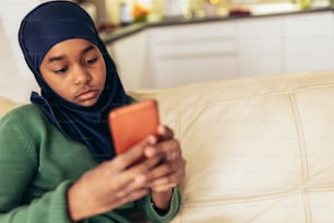 Ragazza musulmana che si siede sul divano a casa usando il telefono.