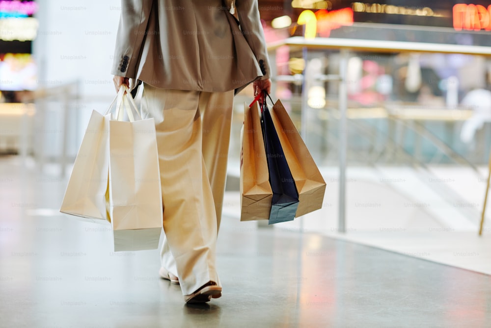 Sección baja de mujer irreconocible con pantalones y sosteniendo bolsas de compras en blanco mientras camina en el centro comercial, copie el espacio