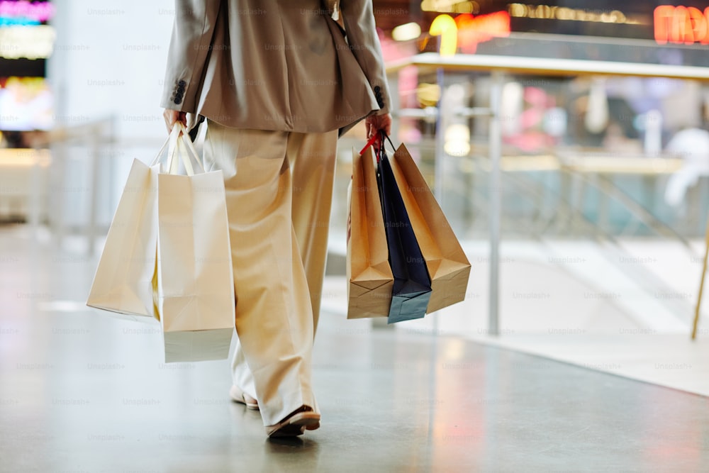 Low section of unrecognizable woman wearing pants and holding blank shopping bags while walking in mall, copy space