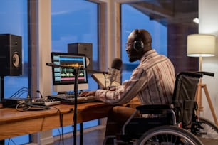 Giovane uomo nero felice in sedia a rotelle che suona la tastiera del pianoforte, canta e registra una nuova canzone in studio o a casa