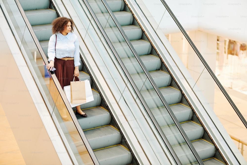 Vista grande angular gráfica em jovem negra descendo escada rolante em shopping center e segurando sacolas, espaço de cópia