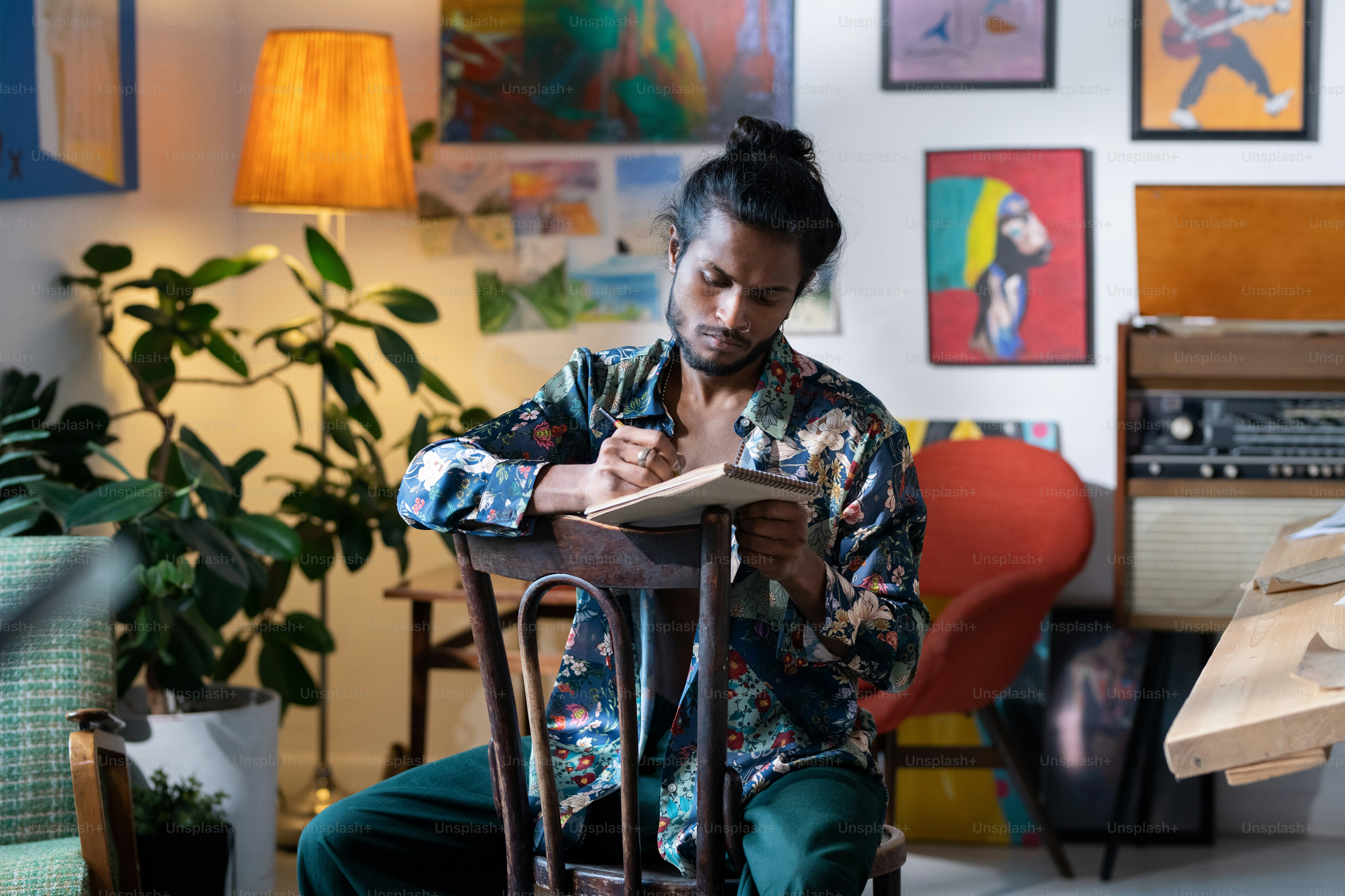 Zeitgenössischer Student der Kunstfakultät zeichnet Skizzen im Notizblock, während er auf einem Stuhl vor der Kamera im Studio oder in der Werkstatt sitzt