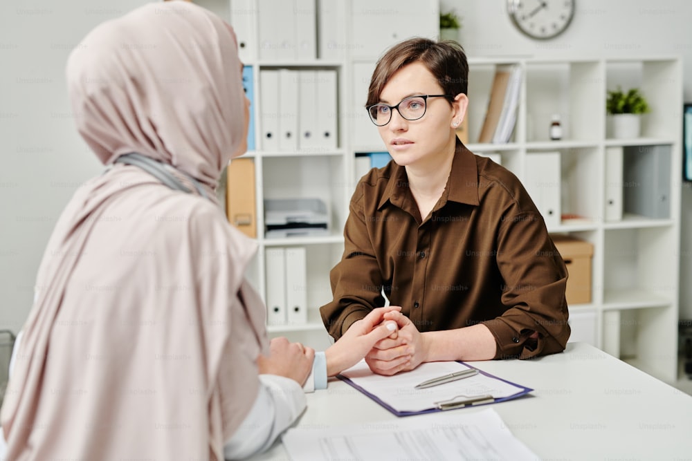 Junge kranke Patientin, die einen muslimischen Arzt im Hijab ansieht, während sie über ihre Diagnose und die Methoden der medizinischen Behandlung spricht