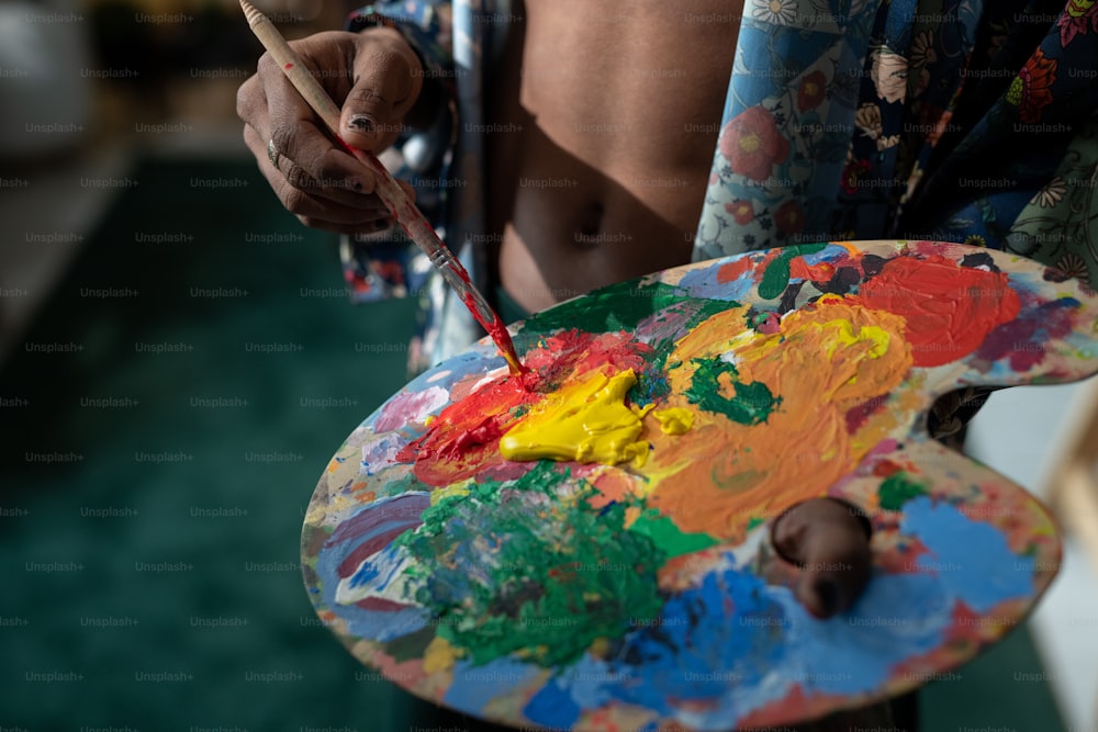 Mani di un giovane uomo non binario con il pennello che mescola colori acrilici sulla tavolozza mentre crea un nuovo capolavoro nello studio d'arte