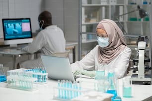 Muslimische Wissenschaftlerin in Hijab und schützender Arbeitskleidung tippt auf der Laptop-Tastatur, während sie am Arbeitsplatz gegen eine Kollegin sitzt