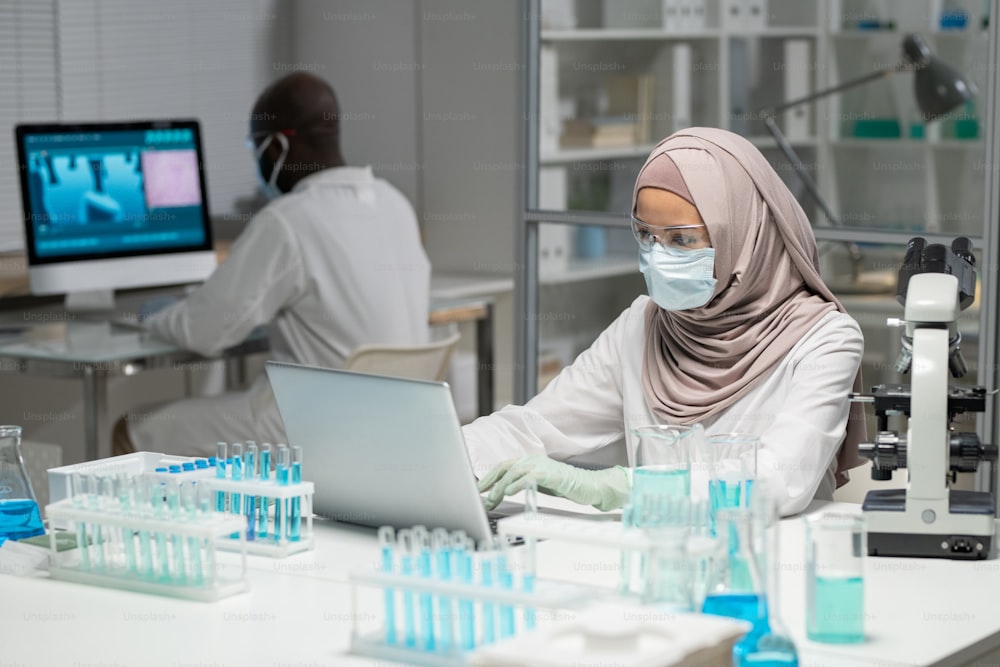 Muslim female scientist in hijab and protective workwear typing on laptop keyboard while sitting by workplace against coworker
