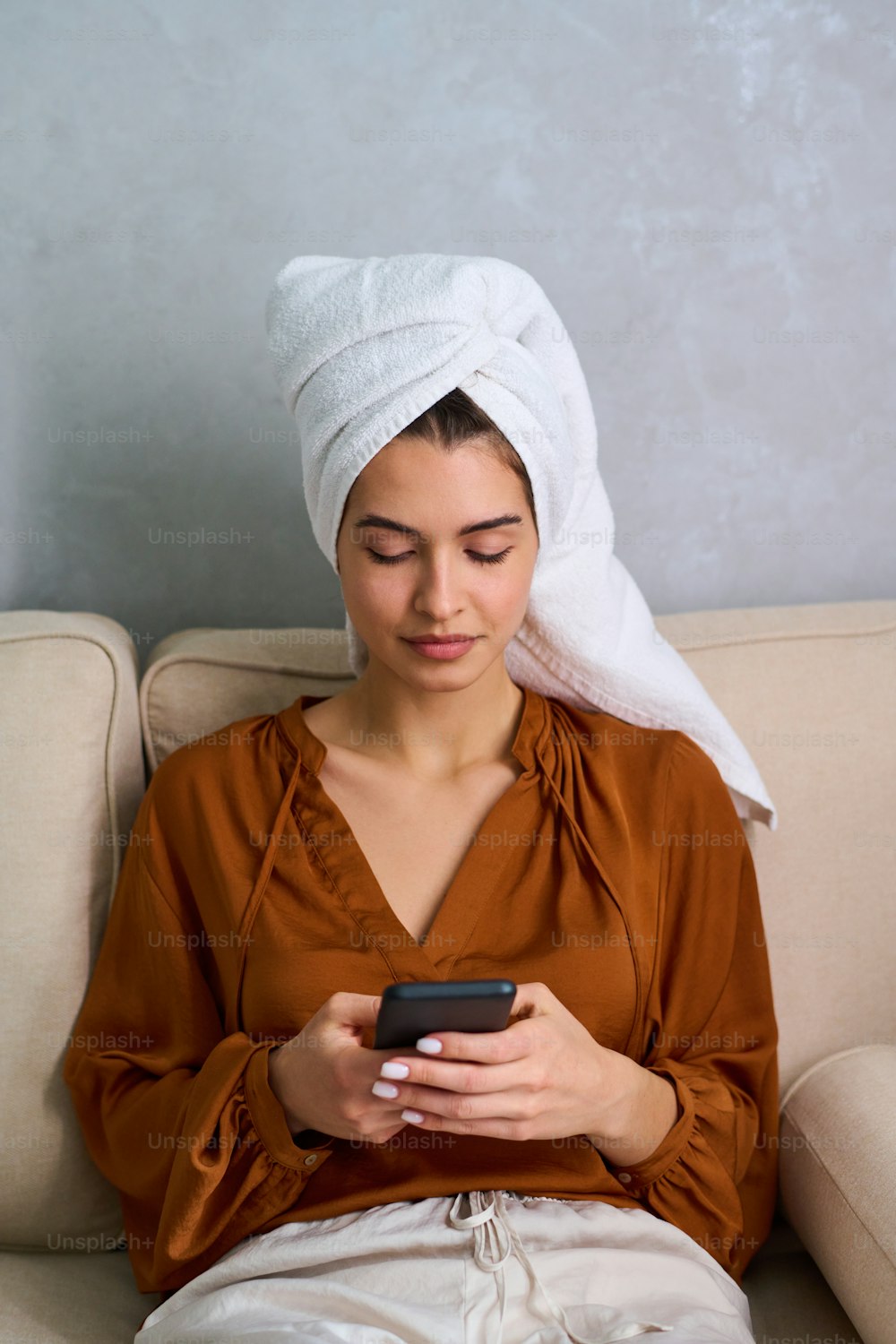 Young serene female in home wear texting in smartphone or communicating in video chat while relaxing on sofa after shower