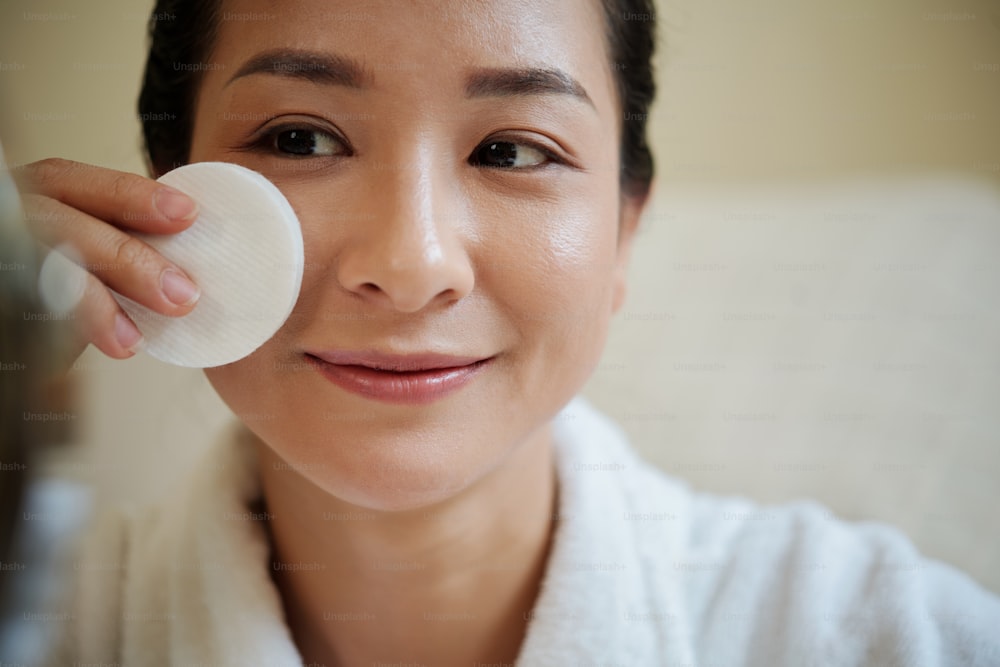 Faccia della donna asiatica sorridente che pulisce il viso con il batuffolo di cotone imbevuto di toner