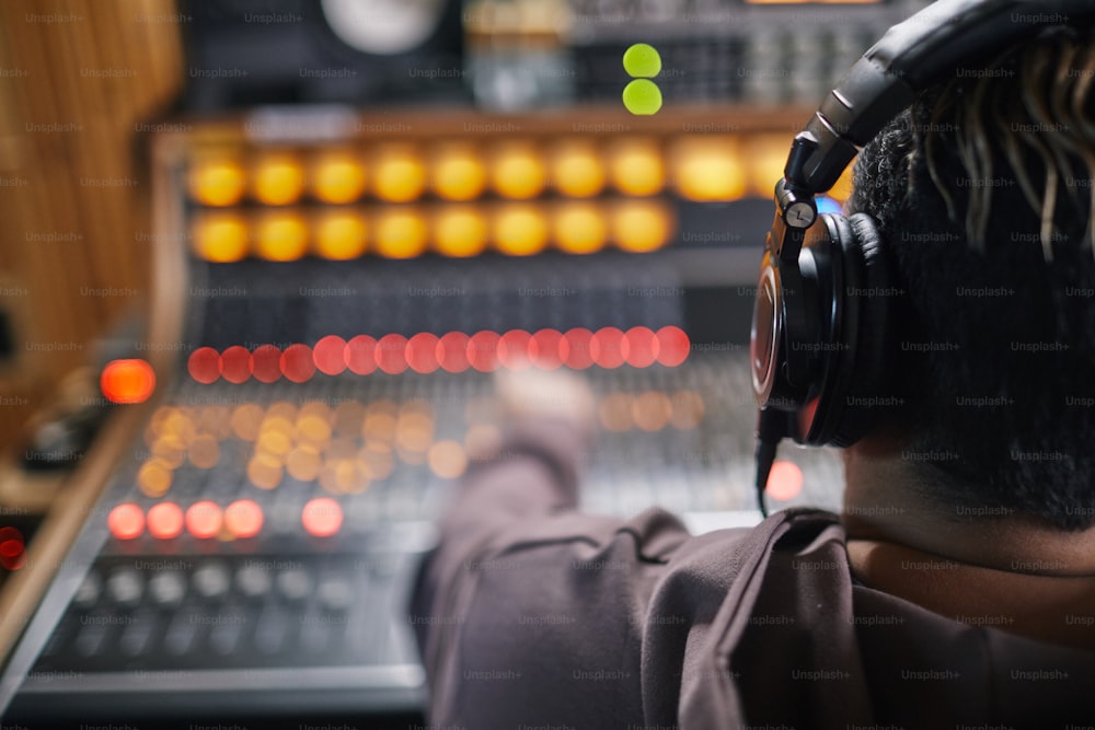 Vista traseira em jovem músico usando fones de ouvido na estação de trabalho de áudio em estúdio de gravação profissional, espaço de cópia